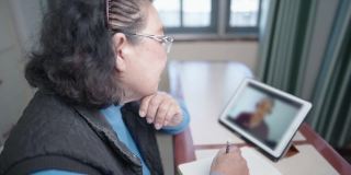 亚洲老妇戴着眼镜看平板电脑在线课程上课，坐下来用铅笔在笔记本上写字，模特穿毛衣，老一代和现代科技退休的生活方式