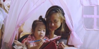 一张专注于亚洲妈妈和可爱的女儿躺在家里粉色帐篷的地板上，在使用数字技术的平板电脑上写着快乐和快乐的时刻。