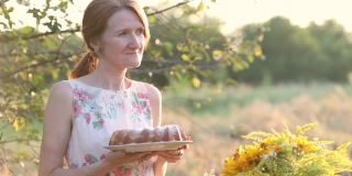 微笑的女人手拿自制的蛋糕站在餐桌旁，在傍晚的阳光照耀下的花园