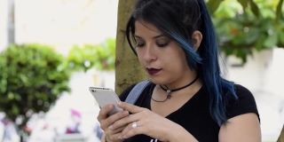 美丽严肃的年轻女子。蓝头发的大学女生在公园的一棵树旁看她的社交媒体。一个女孩在公园里看手机。科技、通讯与教育理念