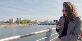 一名女子坐在城市的河边，用她的智能手机在室外打字