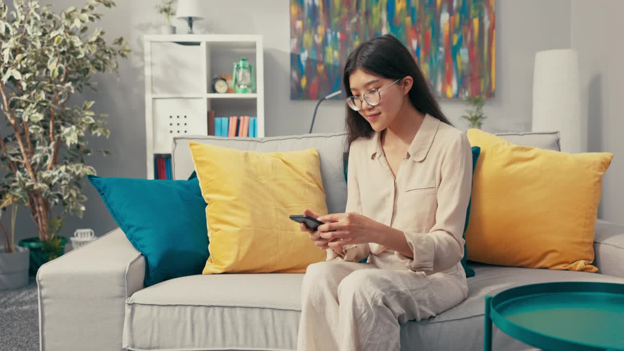 一个美丽的亚洲女人，留着长发，戴着眼镜，坐在客厅的沙发上微笑，穿着亚麻裤和宽松的衬衫，手里拿着手机，和朋友发短信，聊天