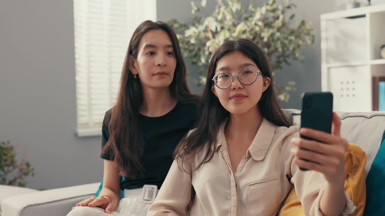 美丽的韩国，亚洲女性在新公寓的沙发上放松，室友，大学生，女士在她面前拿出电话视频通话，交谈，挥手问候，笑容灿烂