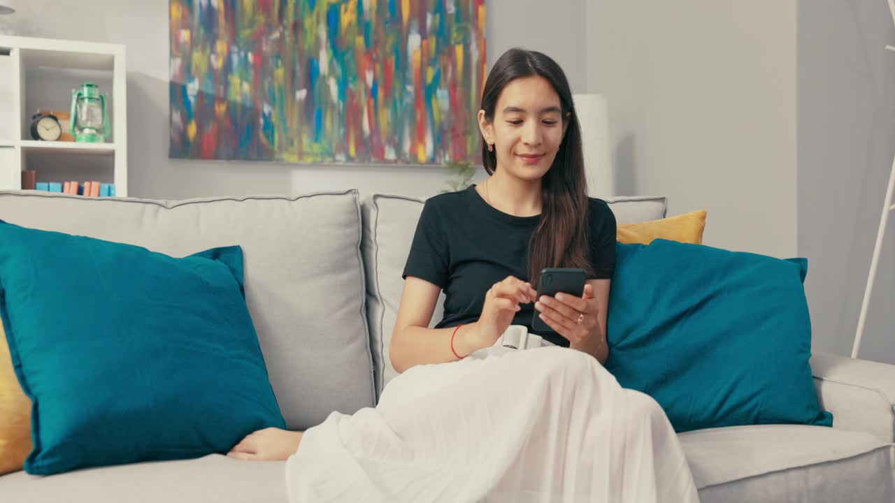 一个美丽的亚洲女人，浅棕色头发坐在客厅的沙发上微笑，穿着亚麻裙子和宽松的黑色t恤，手里拿着手机，与朋友发短信，聊天