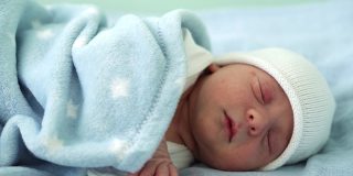 新生儿脸部肖像的特写，在白蓝色的背景上甜蜜地睡觉的早期日子。在帽子上的生命开始分钟的孩子。婴儿，分娩，出生的最初时刻，开始的概念