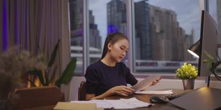 一名亚洲女性坐在她的办公桌前，在家里用电脑和文件工作到深夜。