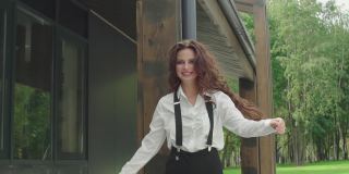 美丽优雅快乐卷曲的女人走在外面。性感的黑头发模特穿着白色衬衫和吊带裤微笑着靠近现代建筑
