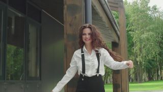 美丽优雅快乐卷曲的女人走在外面。性感的黑头发模特穿着白色衬衫和吊带裤微笑着靠近现代建筑视频素材模板下载
