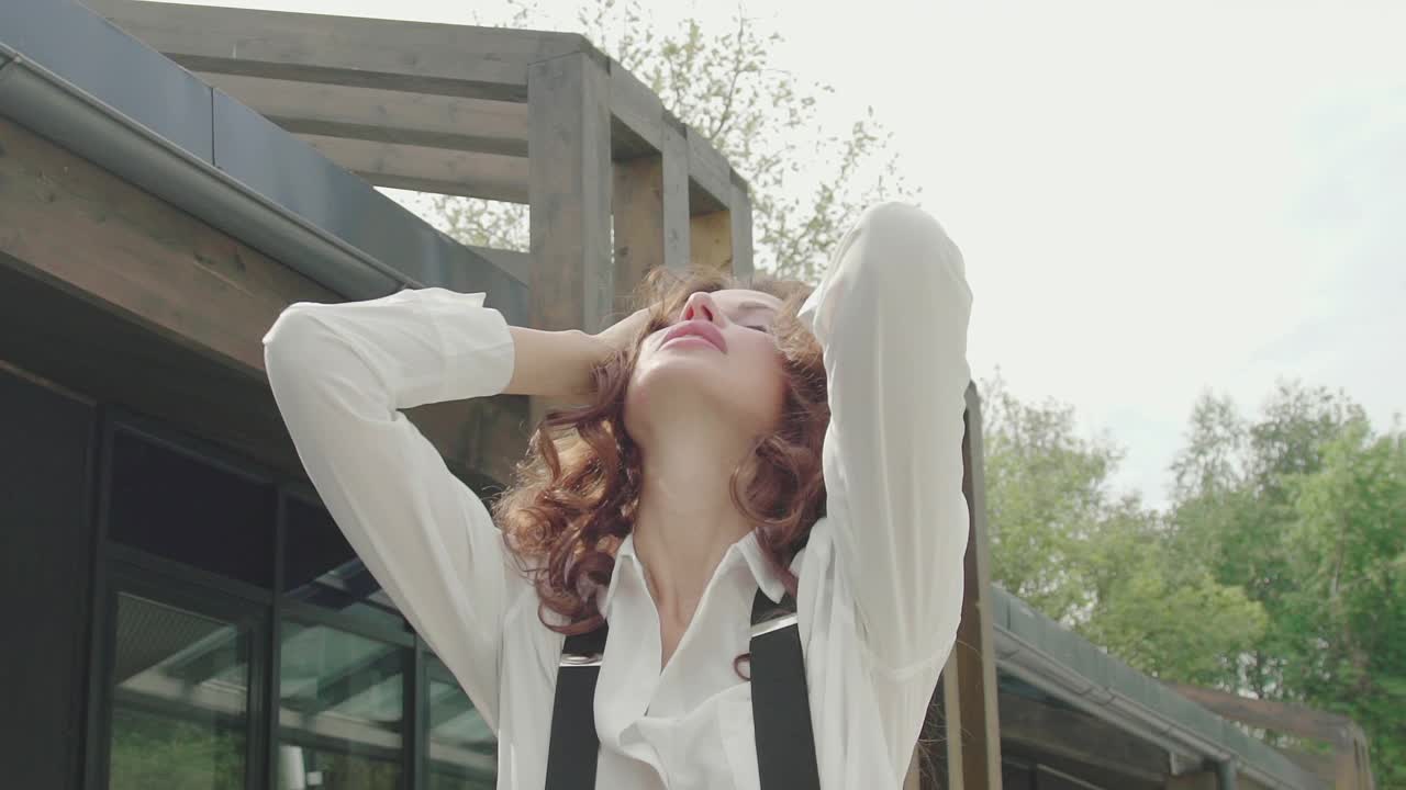 美丽优雅的卷发女人，闭着眼睛享受外面的时刻。性感的黑头发模特穿着白色衬衫和带吊带的裤子在现代建筑附近摆姿势