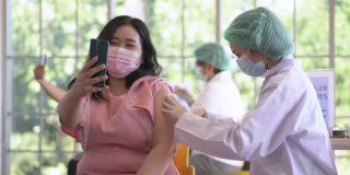 图为一名亚洲女性在诊所或医院接种新冠疫苗时的自拍照片，护工正在注射疫苗以获得保护病毒的免疫力。人们戴着防护口罩。