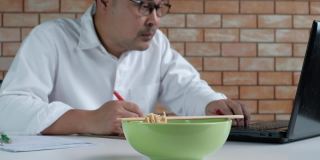 亚洲男性忙于工作，午餐时间在办公室吃方便面。