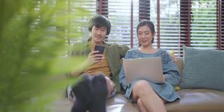 可爱的亚洲结婚夫妇，成年呆在家里的生活方式，男性玩游戏，智能手机，上网，和妻子笑，笑，使用笔记本电脑设备，坐在沙发上，一起放松，客厅在家里，呆在家里的概念