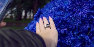 一个女孩用智能手机拍下了她的手，在亮蓝色花朵的背景下，她修剪着漂亮的指甲。带着智能手机和社交网络在公园里散步