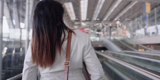 女商人在机场往前走，亚洲女商人，在去机场做生意的时候用智能手机和同事讨论行李和肩袋在自动扶梯上，手持式后面看