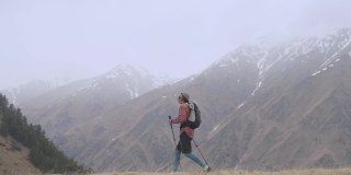 一个年轻人背着双肩包，拿着追踪棒，在低云低岭的多云天气里，爬上了山上的一座小山。徒步旅行的概念