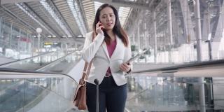 机场商务交流亚洲女商人，在机场出差，带着行李和肩袋在自动扶梯上与顾客交谈，低角度手持前视