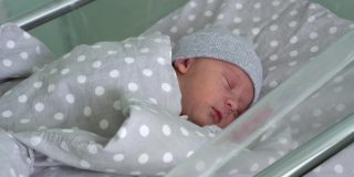 刚出生的婴儿红色可爱的脸肖像早期睡在灰色背景的医用玻璃床上。《帽子上生命开始的几分钟》中的孩子。婴儿，分娩，出生的最初时刻，开始的概念