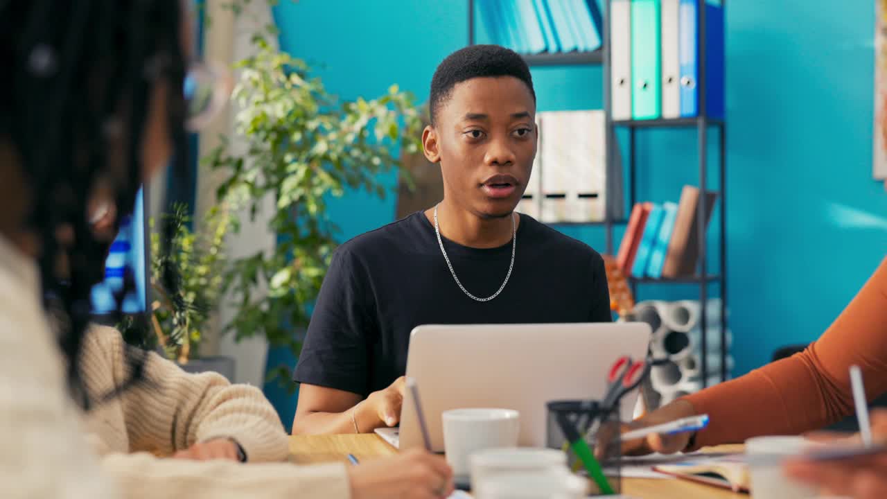 年轻英俊的深色皮肤坐在同事中间，讨论项目的愿景，看着笔记本电脑，谈话，在平板电脑上向每个人展示标识的样本图形