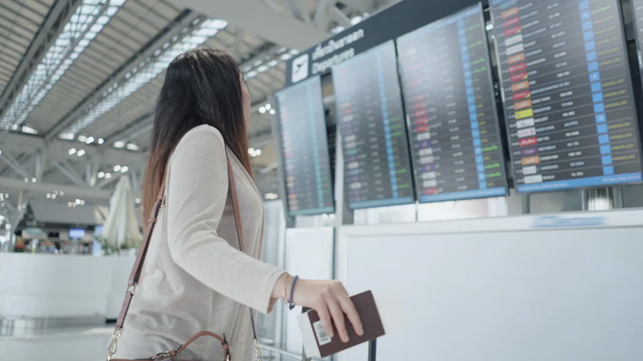 一名亚洲女性旅客在机场，看着她旅行的航班带着行李，在屏幕上看到航班号就开始了新的旅行，旅行度假