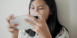 亚洲女孩坐在家里的床上玩手机游戏，生活理念。