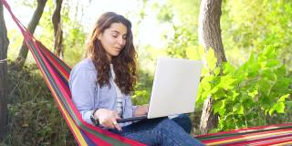 迷人的年轻女子深色头发坐在彩色吊床和使用无线笔记本电脑视频聊天
