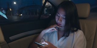 亚洲女商人穿着时尚的办公室服装工作到很晚，在城市现代城市的汽车后座上使用智能手机。