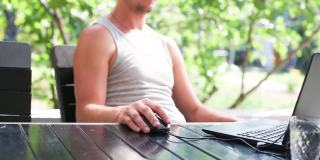 在户外庭院里用笔记本电脑工作——一个穿着t恤的男人坐在桌子旁的野营椅上。家庭办公室，远程工作，远程位置。使用键盘和鼠标，放松
