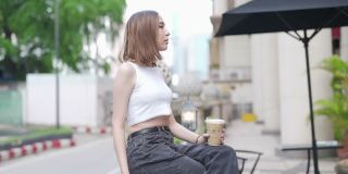 在城市里，年轻的亚洲女人坐在户外长凳上喝着冰咖啡。