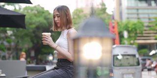 在城市里，年轻的亚洲女人坐在户外长凳上喝着冰咖啡。
