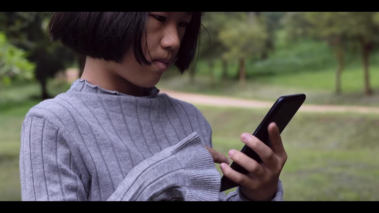 一个亚洲女孩正在室外的绿色公园里看智能手机。4k镜头慢动作