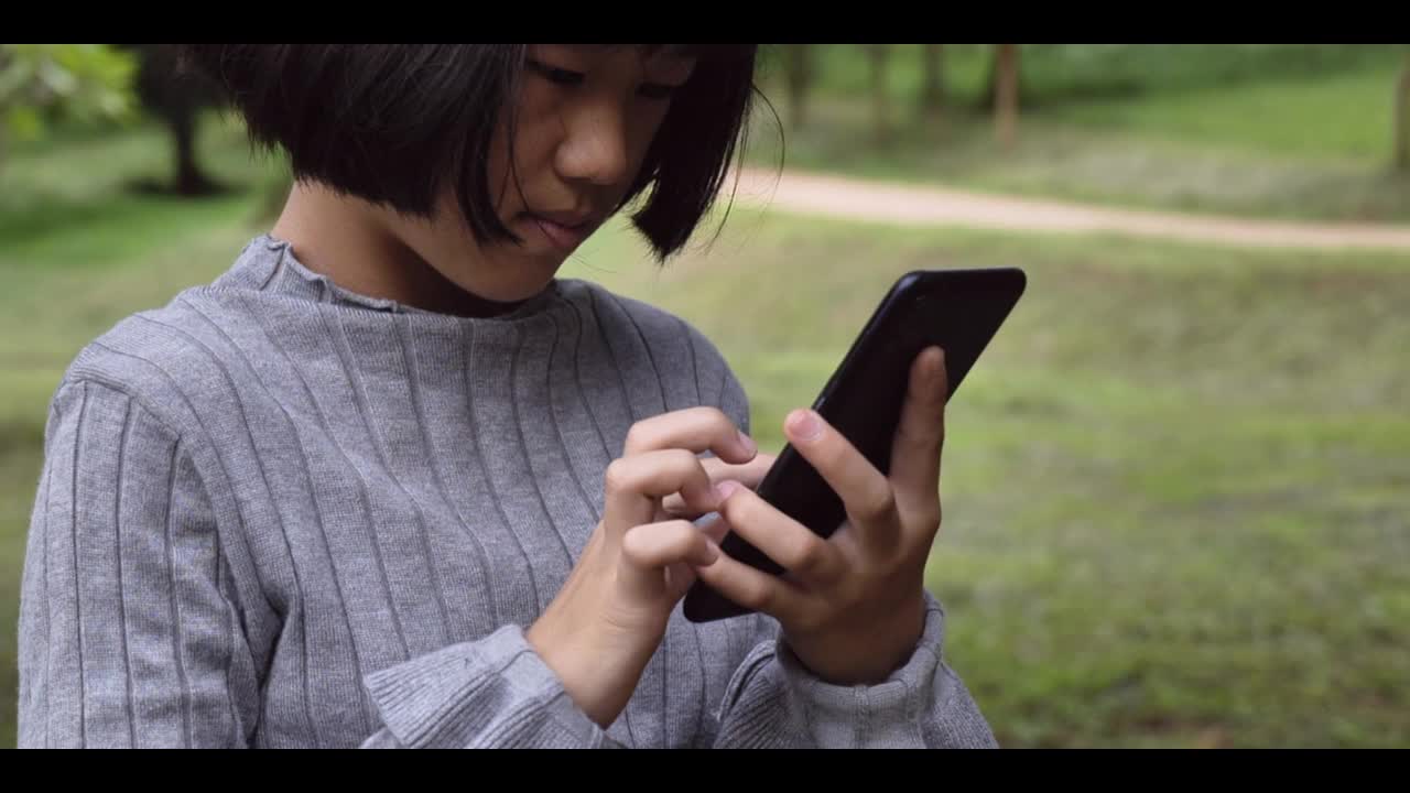 一个亚洲女孩正在室外的绿色公园里看智能手机。4k镜头慢动作