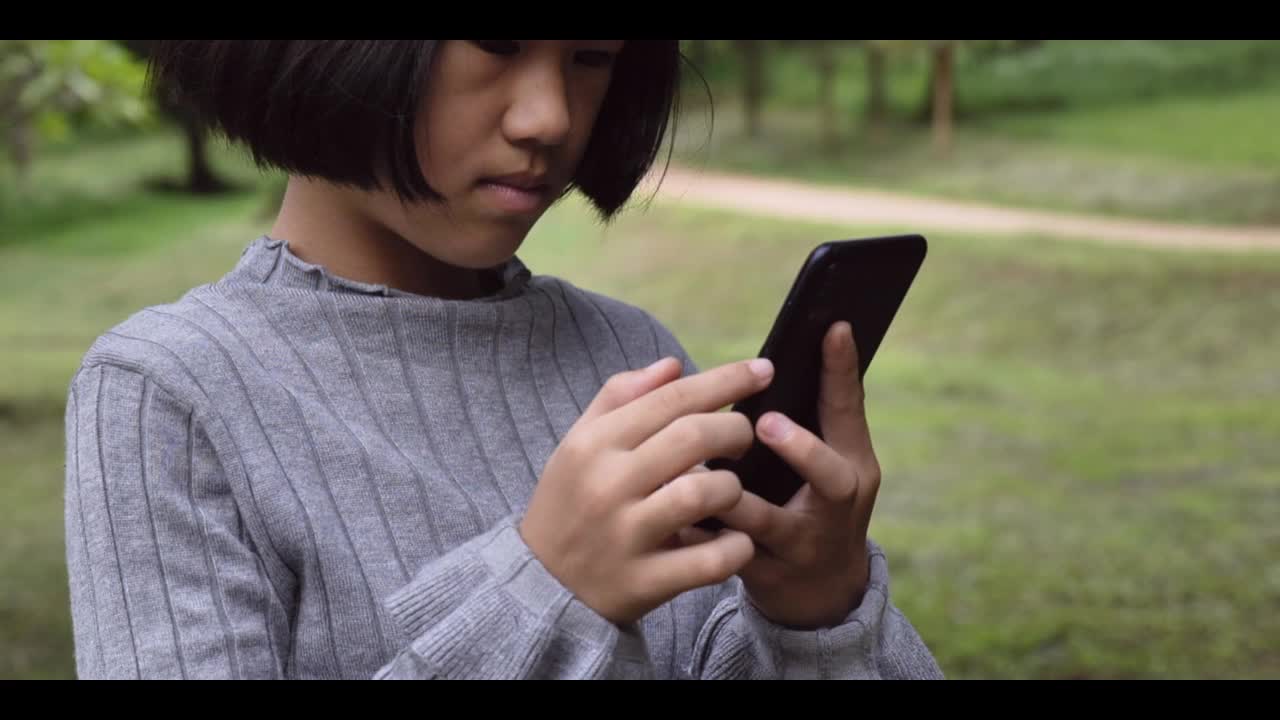 4 k。儿童亚洲女孩看着使用移动智能手机在绿色公园户外。