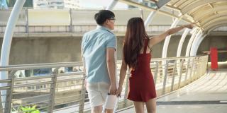 背影背影的年轻亚洲夫妇旅行者与行李在步行和观光在城市交谈。情人旅游者在假期外出旅游。