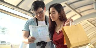 年轻的亚洲夫妇旅行者看地图和指向目的地与行李在城市户外观光。情人旅游和购物袋度假在户外。