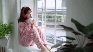 年轻漂亮的亚洲女孩刚刚醒来，在窗边喝着饮料，社交距离概念。视频素材模板下载