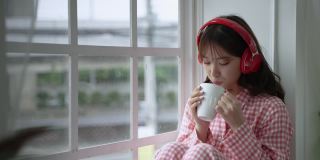 年轻漂亮的亚洲女孩刚刚醒来，在窗边喝着饮料，社交距离概念。