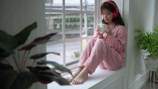 年轻漂亮的亚洲女孩刚刚醒来，在窗边喝着饮料，社交距离概念。视频素材模板下载