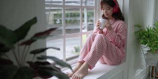 年轻漂亮的亚洲女孩刚刚醒来，在窗边喝着饮料，社交距离概念。