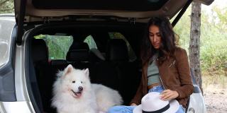 一位戴着时髦帽子的女士带着一只狗坐在一辆车里去大自然旅行。野营的时候,旅行。