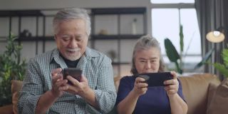 幸福亚洲退休夫妇在客厅家中室内的沙发上一起享受玩游戏和竞争游戏智能手机在线，亚洲夫妇一起玩游戏家隔离的想法