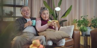 老退休的老亚洲夫妇在家里看电视，老成熟的亚洲夫妇一起在客厅的沙发沙发上欢笑胜利的体育比赛助威活动