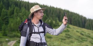 一名真正的白人男性游客的特写照片，他背着背包，拿着手杖在森林附近的一座山上行走，打视频电话，与亲戚和朋友交流。