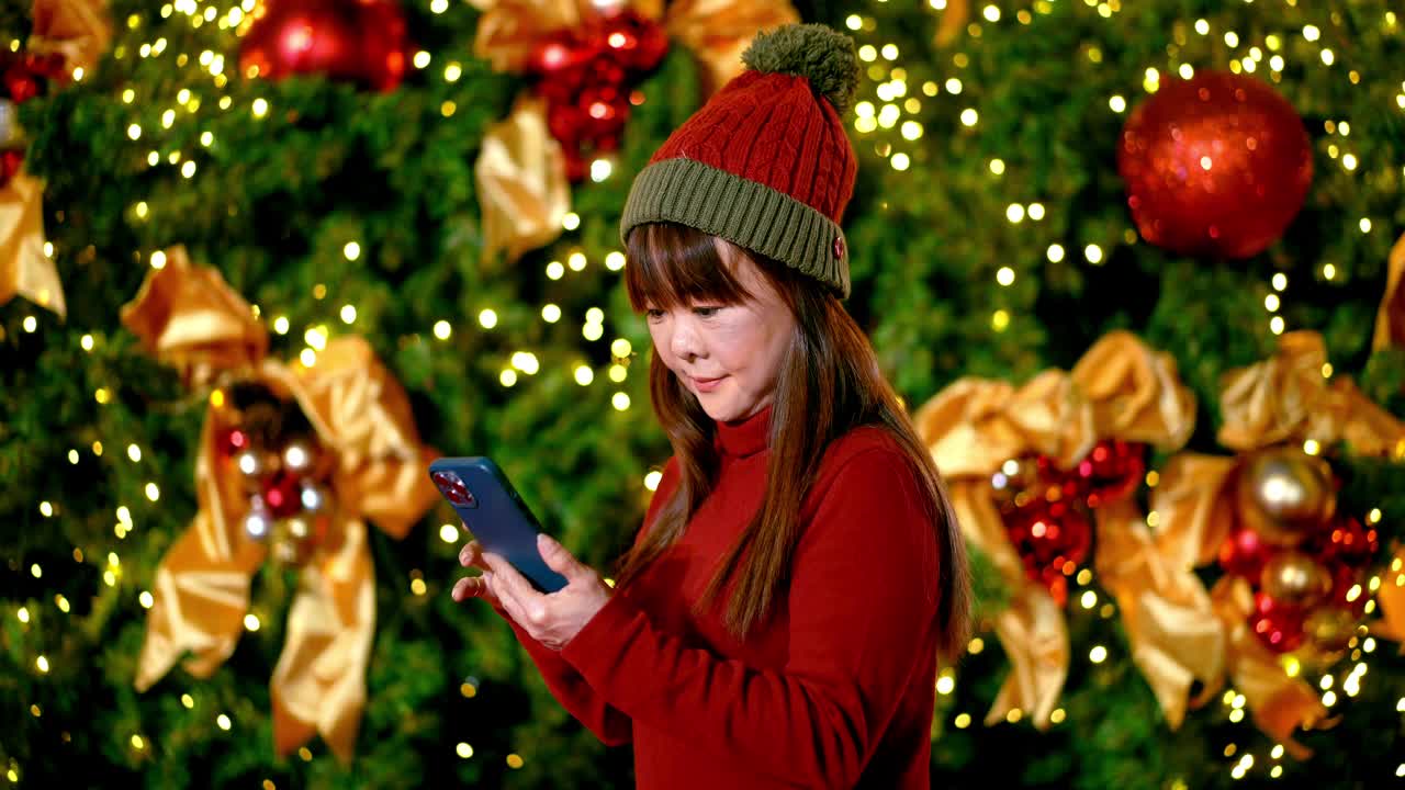穿着红色毛衣在圣诞树前玩手机的成熟女人