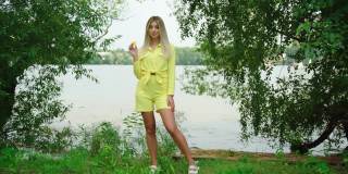 美丽的金发微笑女人享受暑假在池塘。模特穿着黄色的衣服，拿着一片黄色的西瓜。漂亮女孩吃新鲜食物的肖像。