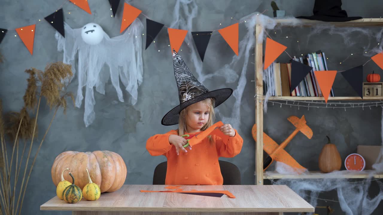 万圣节工艺品。一个戴着女巫帽的白人女孩坐在家里的桌子旁，用剪刀把南瓜和鬼魂剪下来。