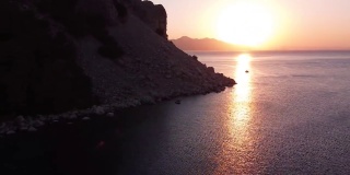 航拍的彩色日落海面与帆船。反射在水面上的太阳。日出的海洋与岩石和山脉。海景，旅游度假的概念。无人机视频