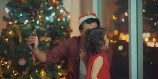 父亲和女儿与他们的家人视频通话，靠近圣诞树和客厅墙上的玻璃在圣诞节，4K分辨率。
