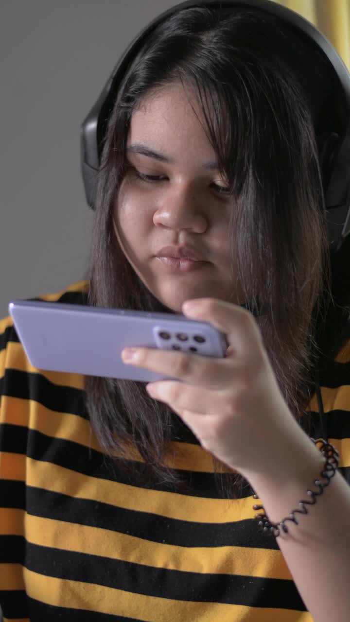 直立肖像亚洲胖脸少女喜欢在家里吃零食的同时戴着耳机玩手机游戏。