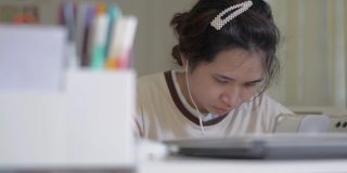 亚洲漂亮的女大学生在休闲服装学习在线上的数字设备与耳机在家里的桌子上。一名泰国裔的年轻女性在研究数字平板电脑。