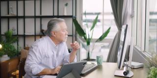亚洲资深成熟男子在家里的桌子上用电脑和平板电脑工作，在忙碌中与客户交谈。